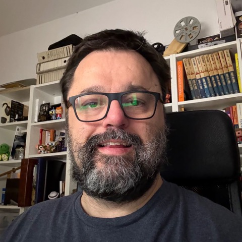 Mariano Cuesta, sonriente, con gafas y barba entrecana, delante de una estantería con libros, cámaras y una bobina de cine