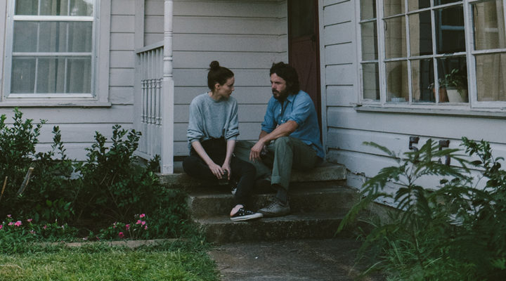 Una pareja joven sentada en los escalones del porche de una casa de madera blanca, mirándose con gesto serio en un ambiente íntimo y melancólico.