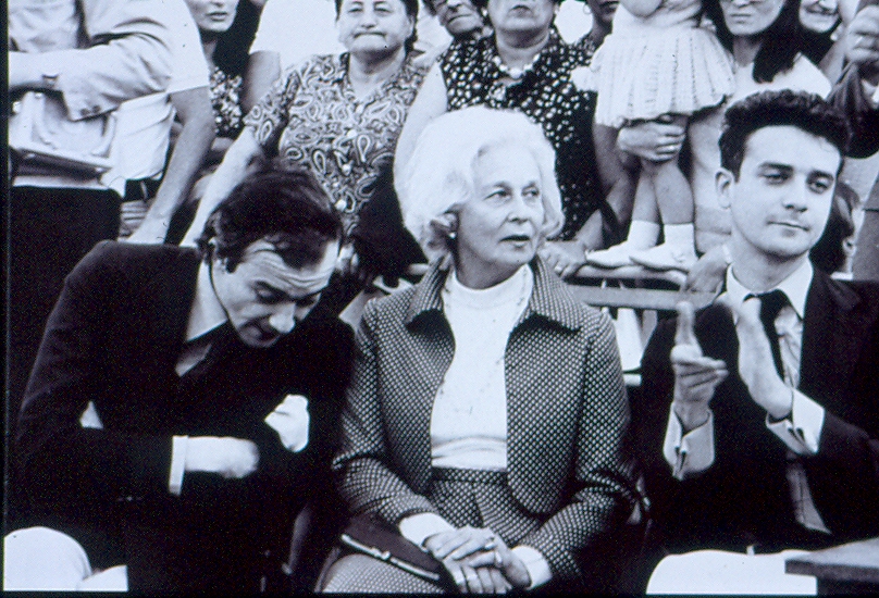 Retrato en blanco y negro de una mujer mayor sentada entre dos hombres jóvenes en una grada abarrotada, mientras el hombre de la derecha aplaude y varias personas observan al fondo.